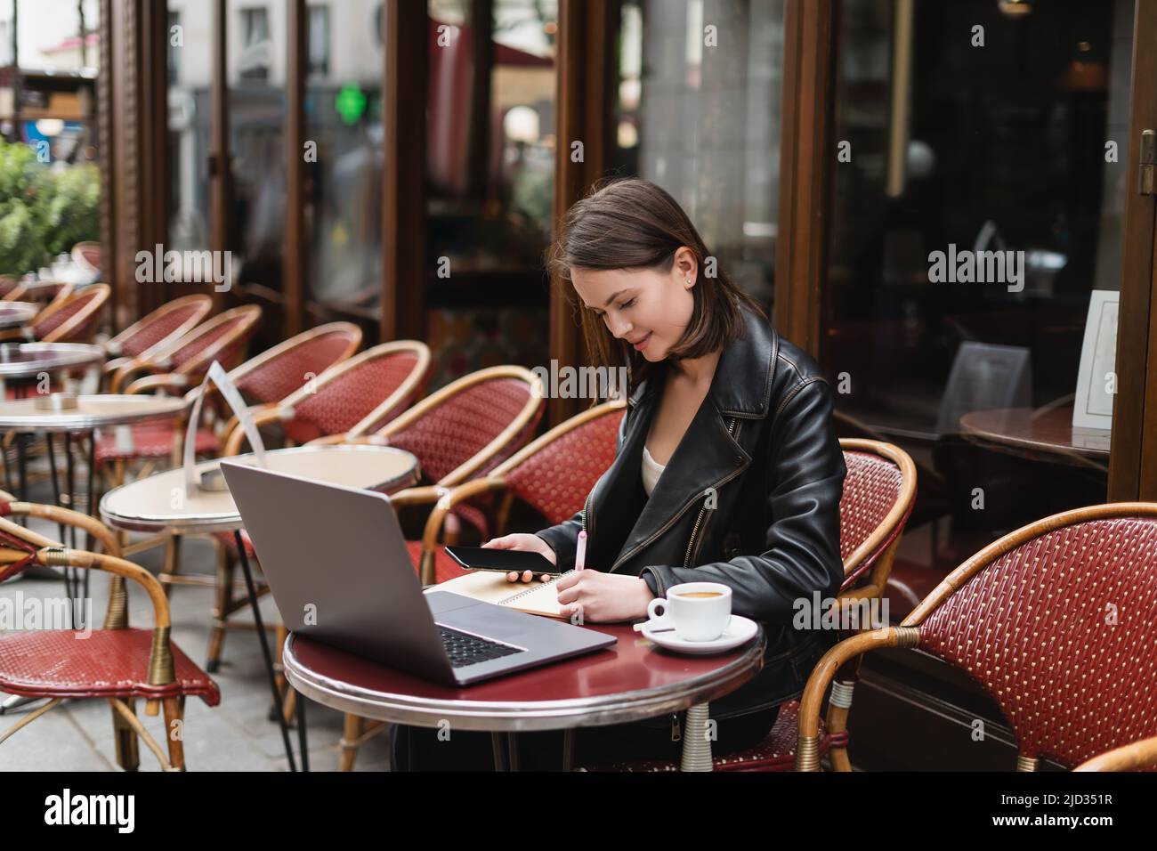 young freelancer in black jacket holding smartphone and writing near laptop and cup of coffee in french outdoor cafe Stock Photo