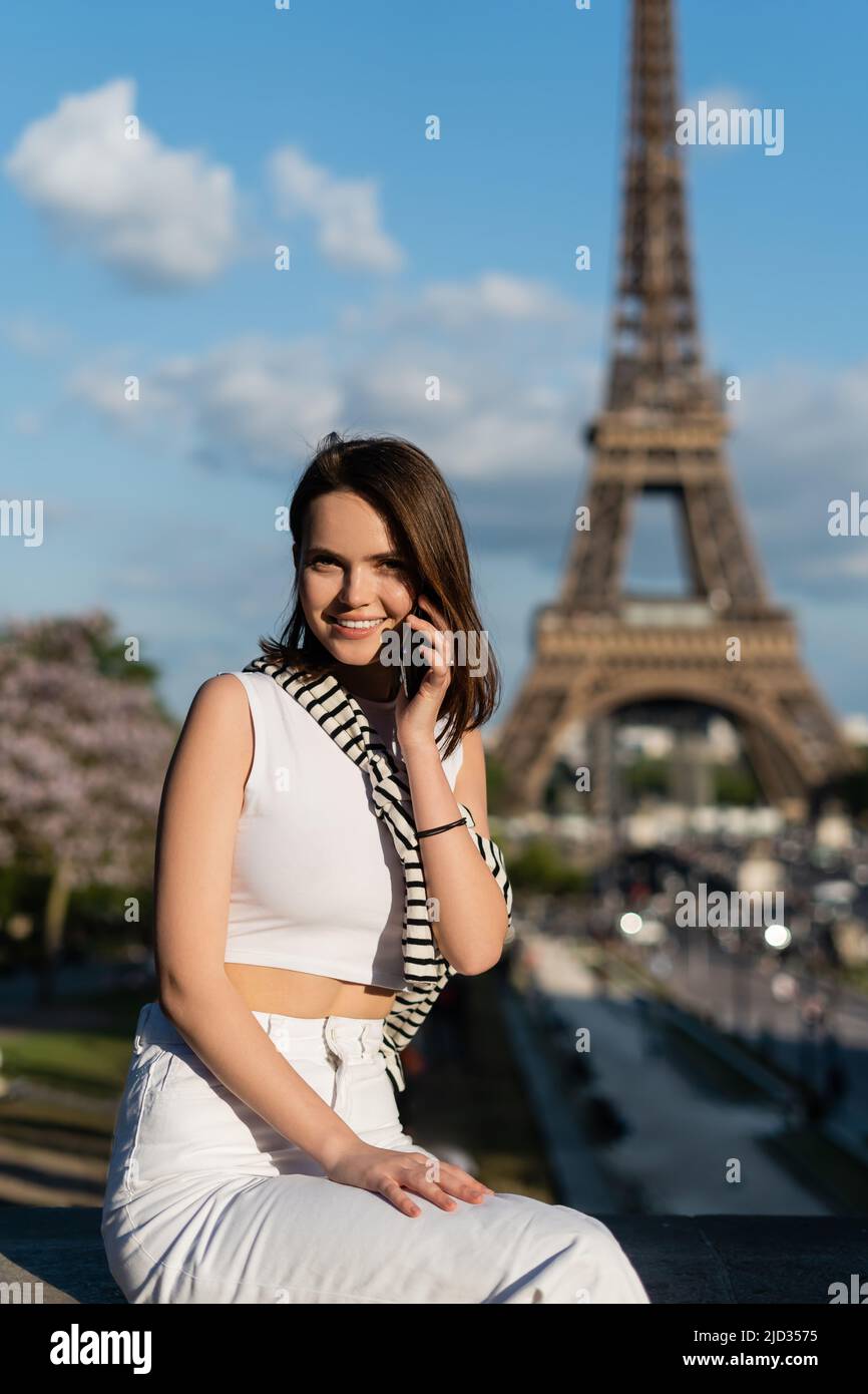 happy young woman in trendy outfit talking on smartphone while sitting near eiffel tower in paris Stock Photo