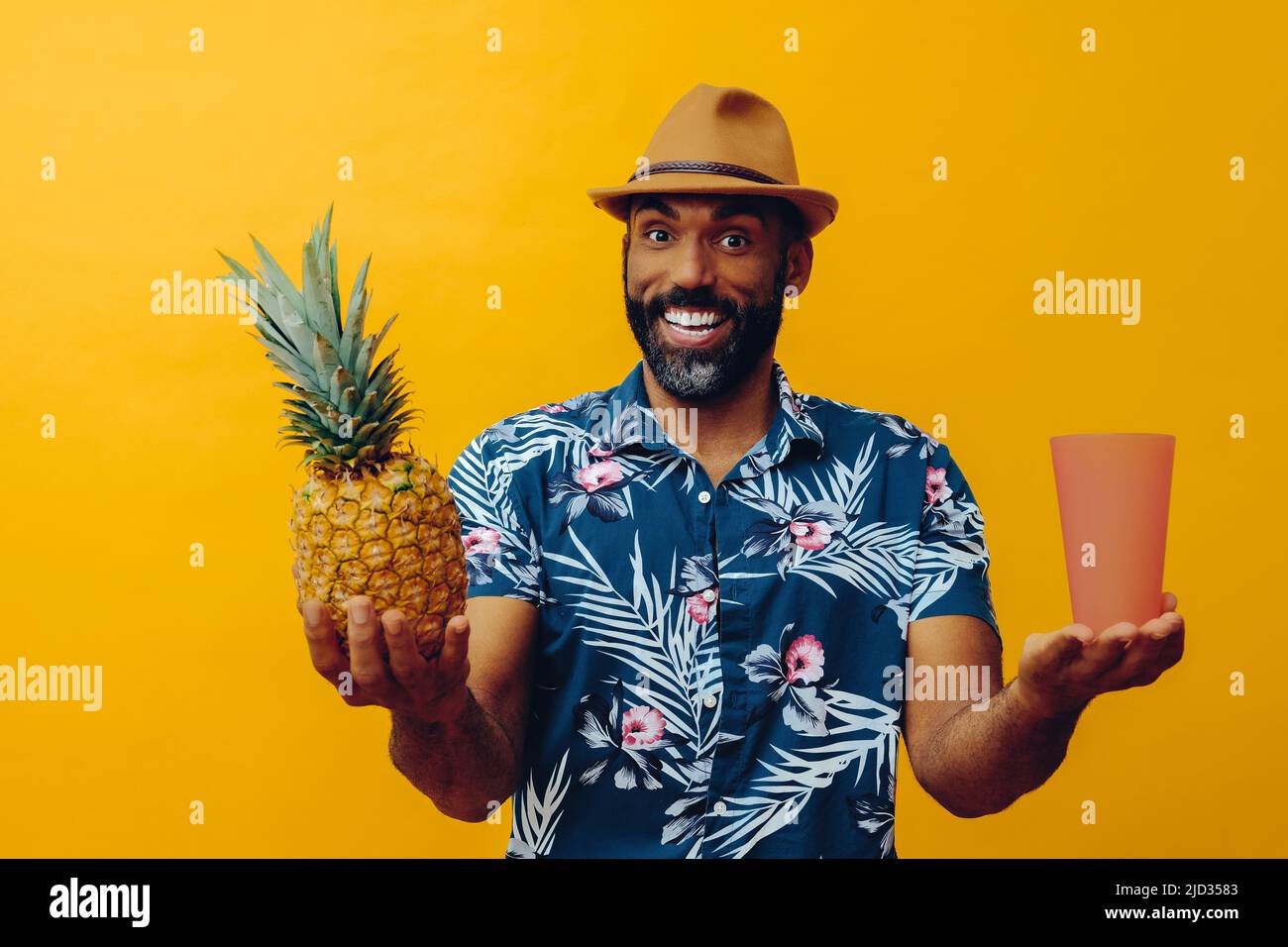 handsome mid adult smiling african american man wearing Hawaiian shirt and hat balancing pineapple and glass with hands to the sides studio shots Stock Photo