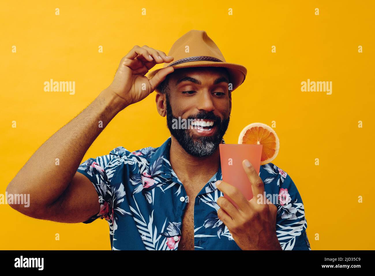 mid adult bearded african american man wearing Hawaiian shirt and hand on hat looking at orange cocktail juice smiling studio Stock Photo