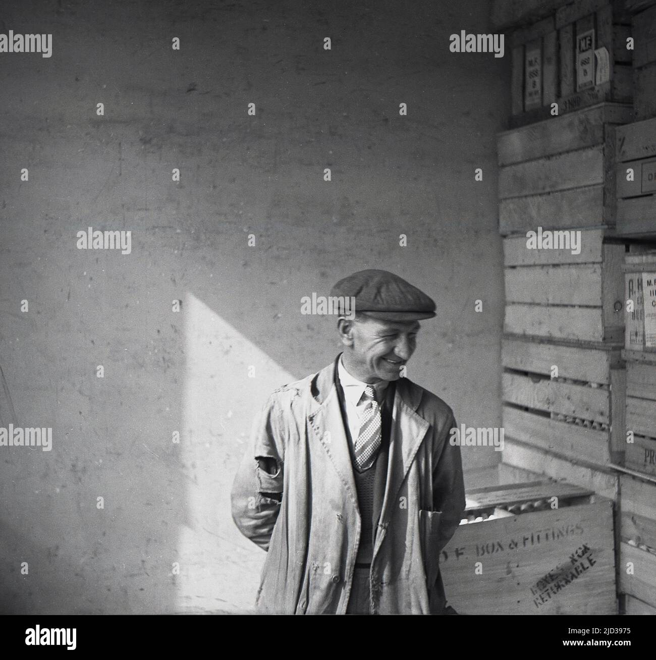 1940s, historical, a male shop/warehouse worker smiling, wearing shirt and tie underneath tatty work overall or coat - full of holes - and a cloth cap, standing for a photo inside, beside a stack of wooden crates, Bramshall, Stockport, Manchester, England, UK. Stock Photo