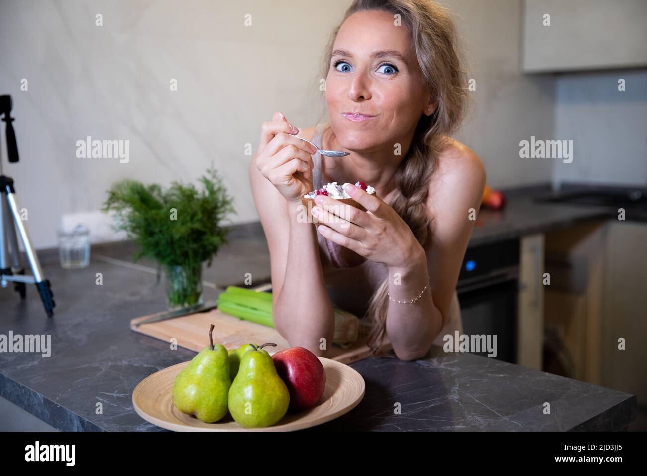 A young beautiful blonde with blue eyes of Caucasian appearance eats with great appetite fresh healthy food at the kitchen table at home. On the face Stock Photo