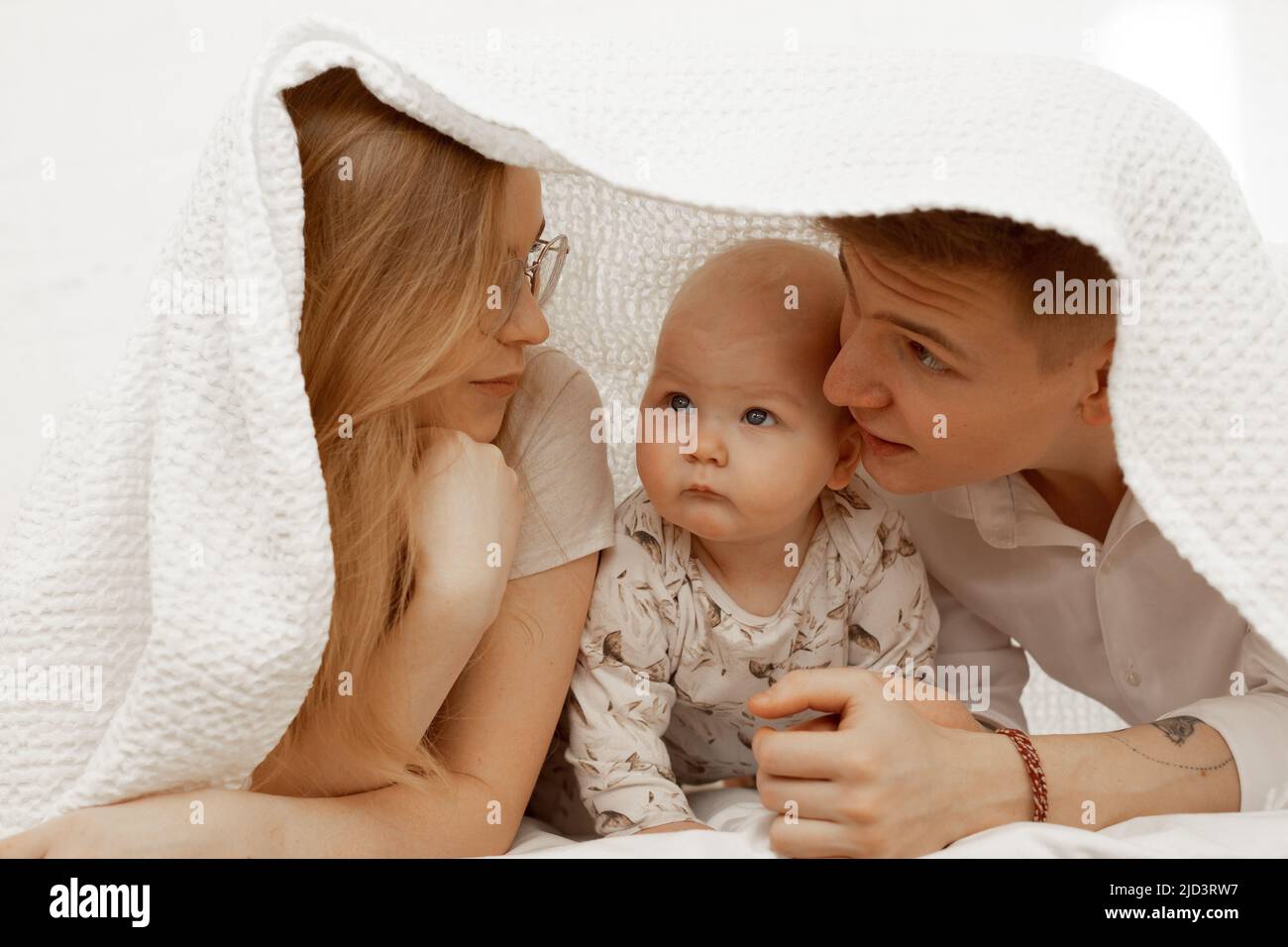 Young mother and father lay under white blanket with little baby on bed and enjoy. Cute relationship between parents and infant child closeup, free Stock Photo