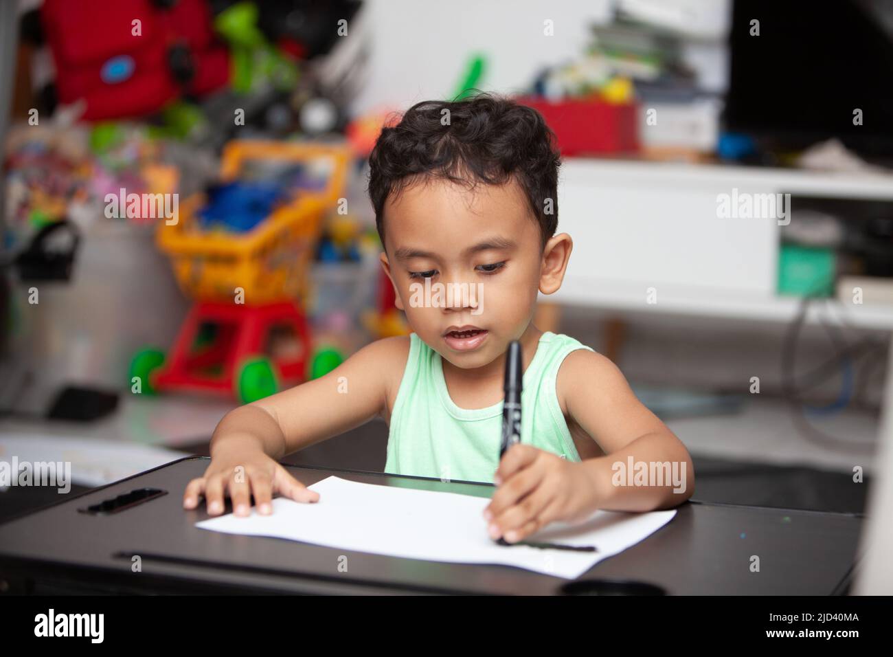 An Indonesian young boy in a light green t-shirt holding a black cryon to color a white sheet of paper on a black table in the living room Stock Photo