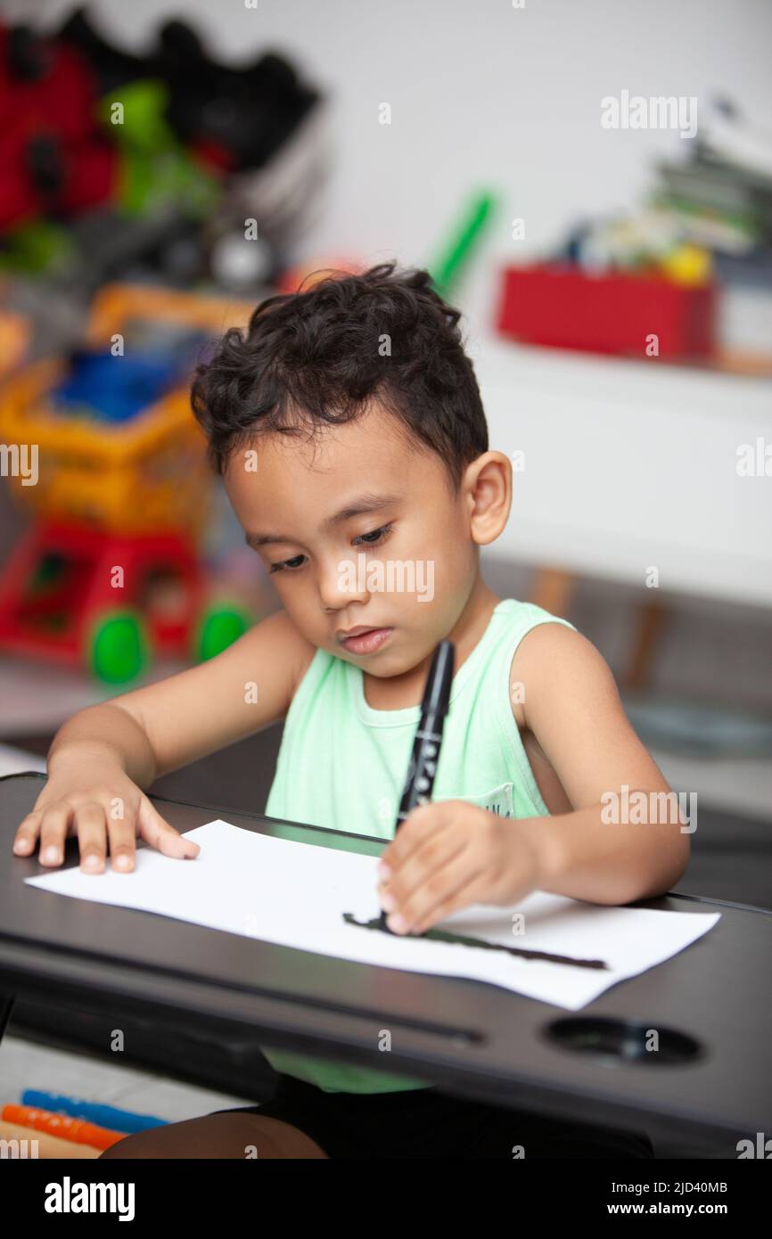 An Indonesian young boy in a light green t-shirt holding a black cryon to color a white sheet of paper on a black table in the living room Stock Photo