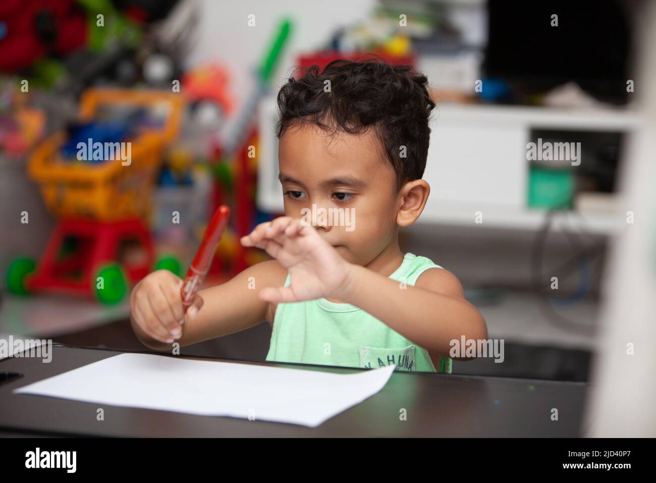 An Indonesian young boy in a light green t-shirt holding a red crayon to color a white sheet of paper on a black table in the living room Stock Photo