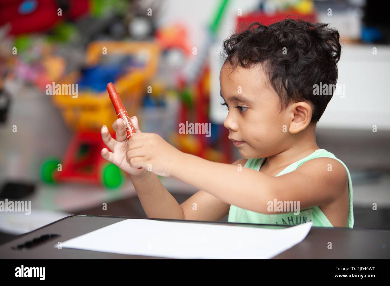 An Indonesian young boy in a light green t-shirt holding a red crayon to color a white sheet of paper on a black table in the living room Stock Photo