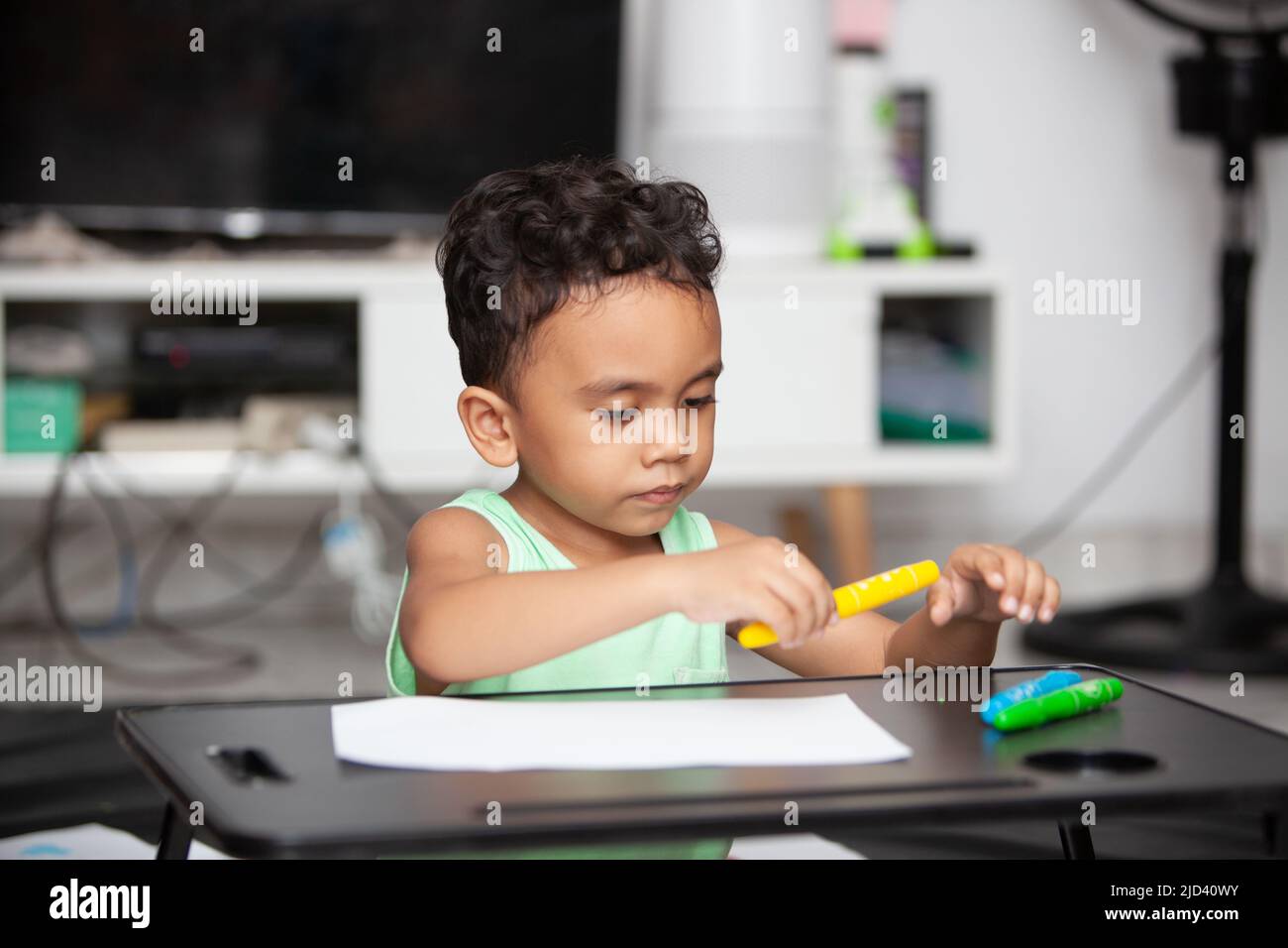 An Indonesian young boy in a light green t-shirt holding a yellow crayon to color a white sheet of paper on a black table in the living room Stock Photo