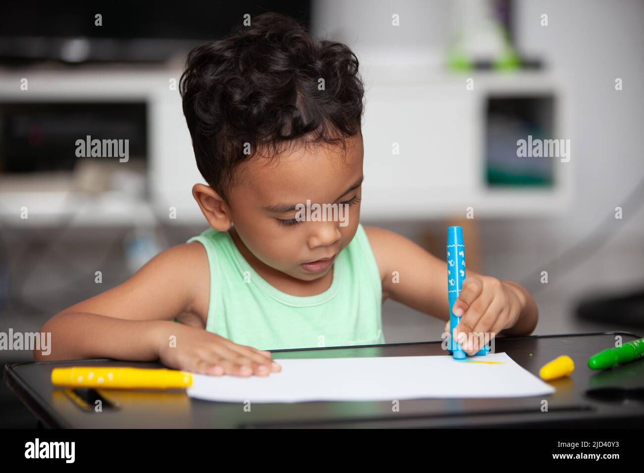 An Indonesian young boy in a light green t-shirt holding a blue crayon to color a white sheet of paper on a black table in the living room Stock Photo