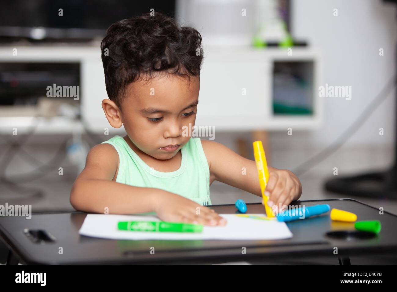 An Indonesian young boy in a light green t-shirt holding a yellow crayon to color a white sheet of paper on a black table in the living room Stock Photo