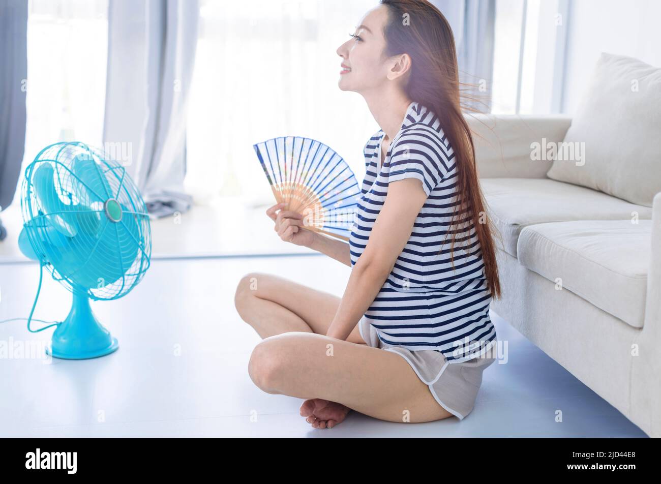 Young woman relax on floot in living room wave with hand fan and enjoying air flow from fan . Summer heat concept Stock Photo