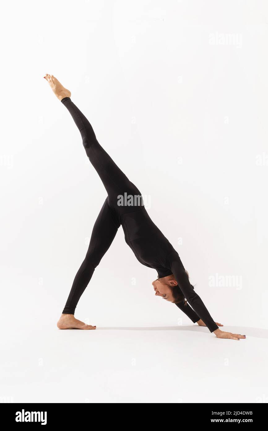 A female yoga practitioner doing a variation of the Adho Mukha Svanasana exercise, downward facing dog pose, exercising in a black bodysuit on a white Stock Photo