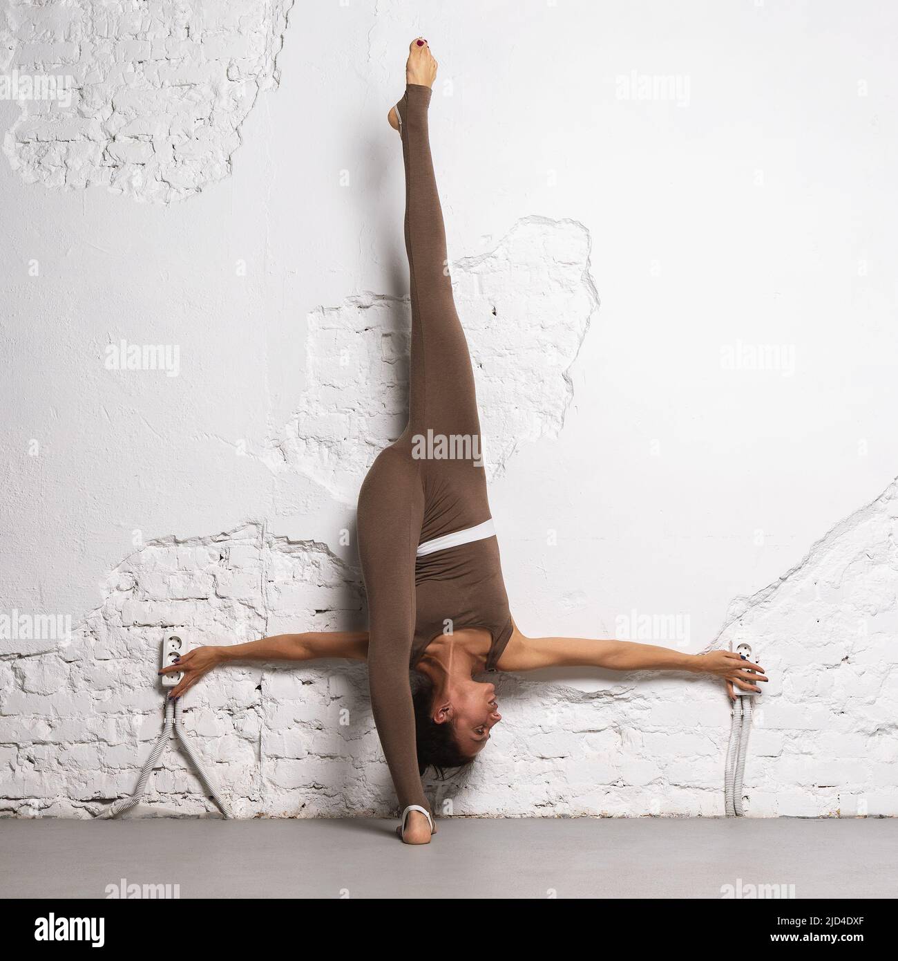 A woman is engaged in stretching the muscles of her legs, performs a longitudinal split, leaning her back against the wall, and trains in the room Stock Photo