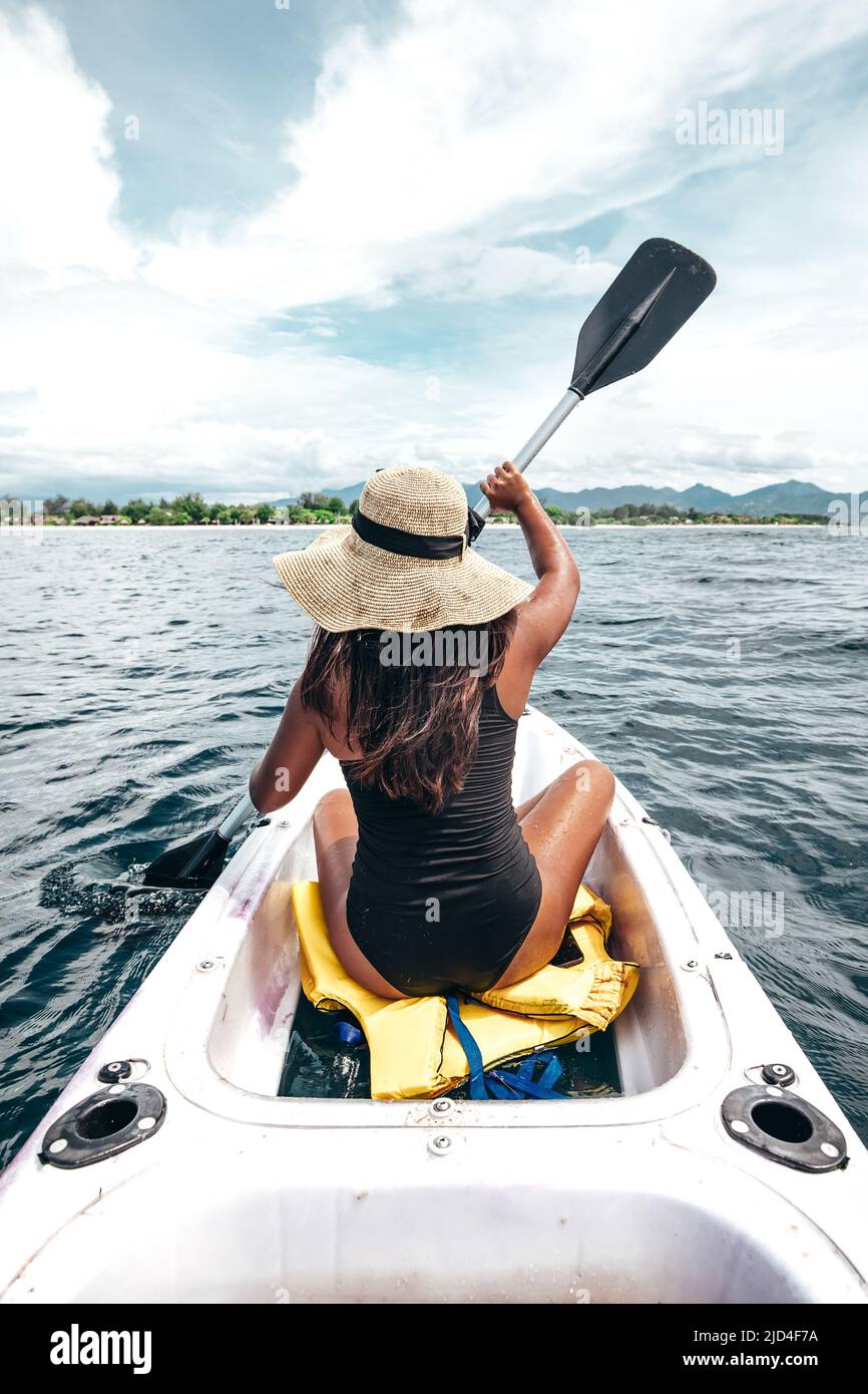 young Indonesian woman in black swimsuit and hat paddling in a white