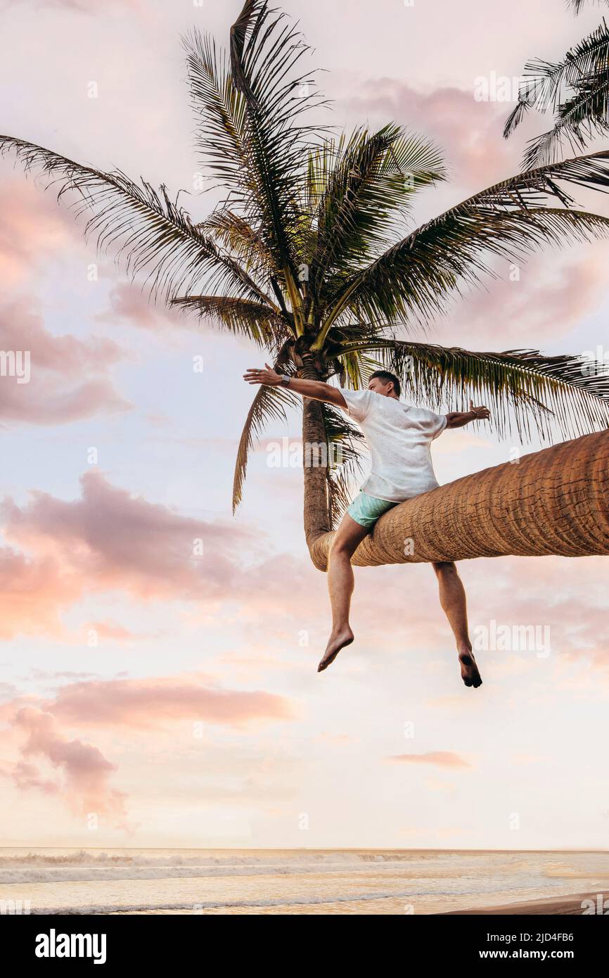 excited young man siting on curved coconut tree at a black sand beach in Bali during sunset