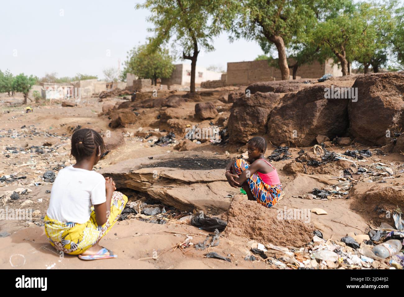 Two sad African village girls crouch on the dusty and garbage-filled roadside, among stones, sand and sparse vegetation, symbolizing the decay and des Stock Photo