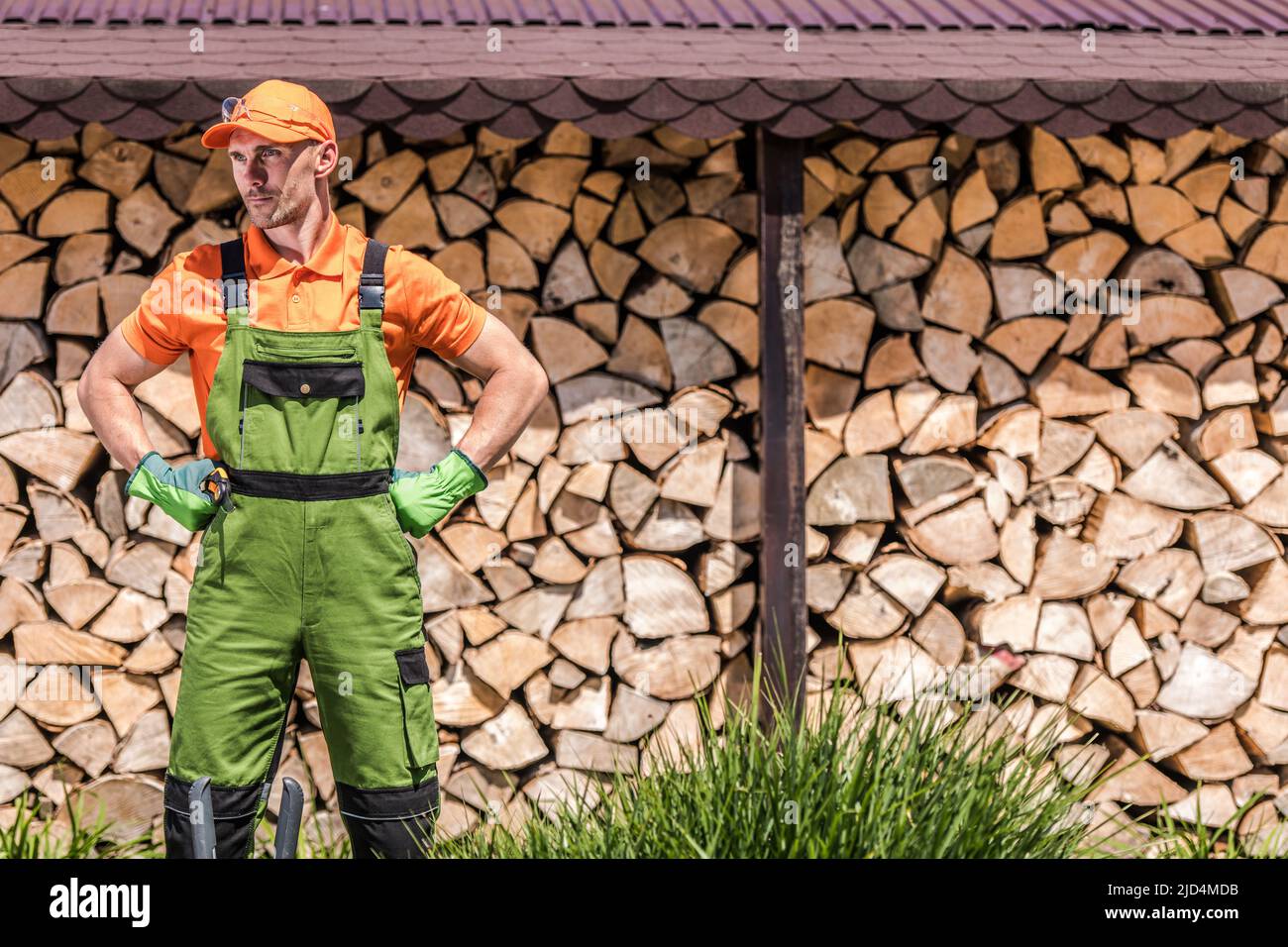 Male Caucasian Worked Dressed in Professional Gardening Wear Standing in Front of Firewood Wall Made of Chopped Wood Pieces. Stock Photo