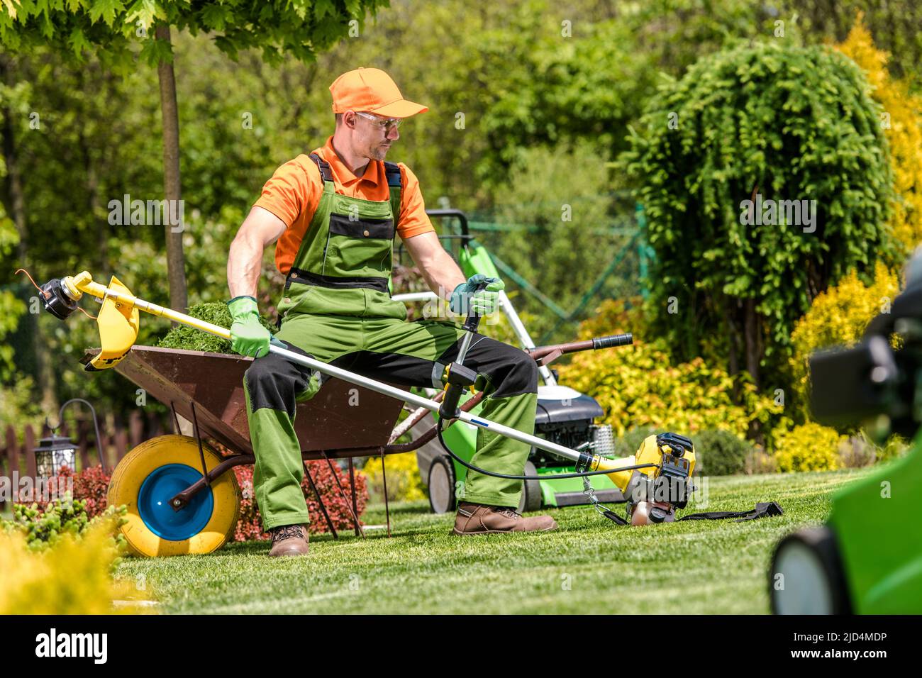 Caucasian Landscape Gardener Resting While Mowing the Lawn Sitting on the Wheelbarrow and Holding Professional Grass Trimmer in His Hands. Stock Photo