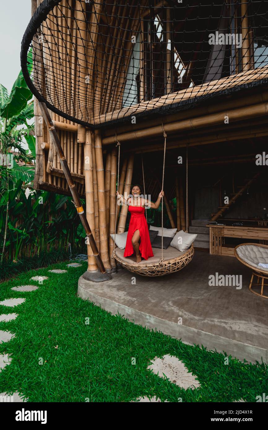 young happy Indonesian woman in red maxi dress smiling at luxury bamboo eco villa in ubud bali Stock Photo