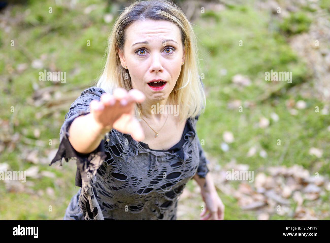 Wild angry blond woman reaching up to claw at the camera with a snarl as she crouches beside a forest tree Stock Photo