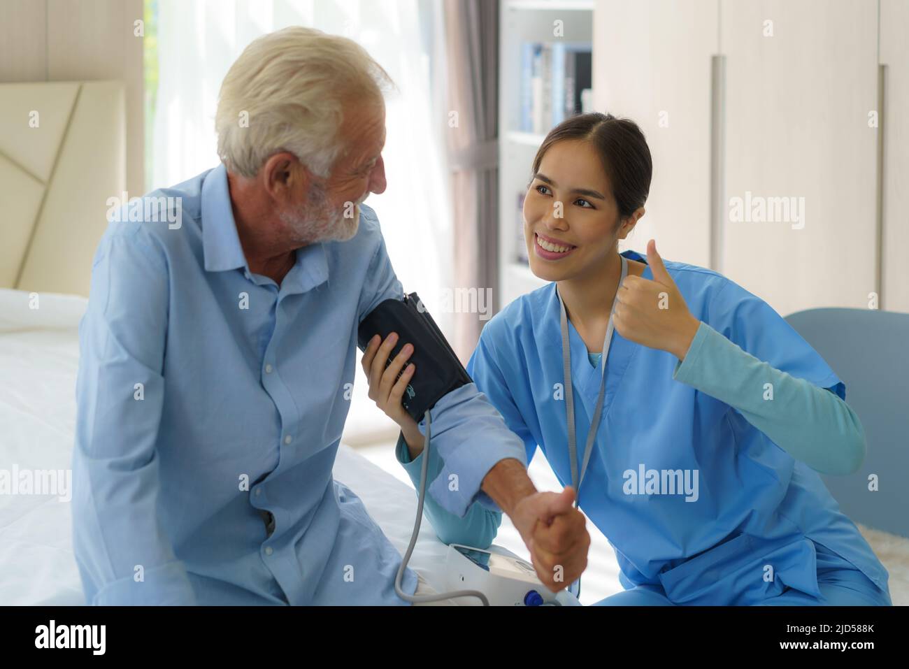 Asian Attentive caregiver using tonometer while elder man having breath problem. Female doctor measuring blood pressure of senior man at home. Stock Photo