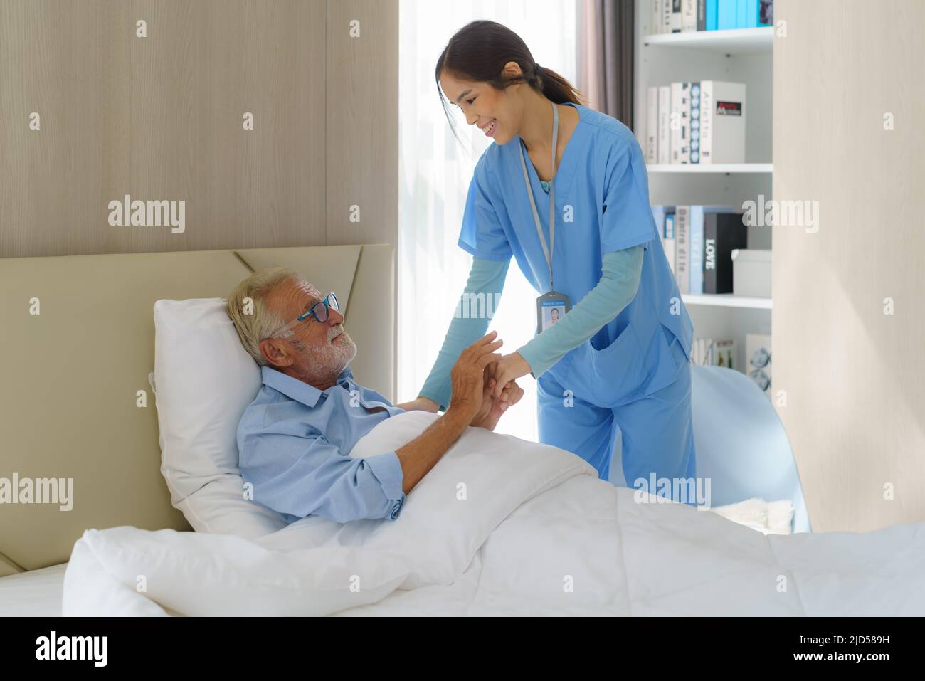 Asian nurse standing on a home bed next to an older man helping hands, care. Elderly patient care and health lifestyle, medical concept. Stock Photo