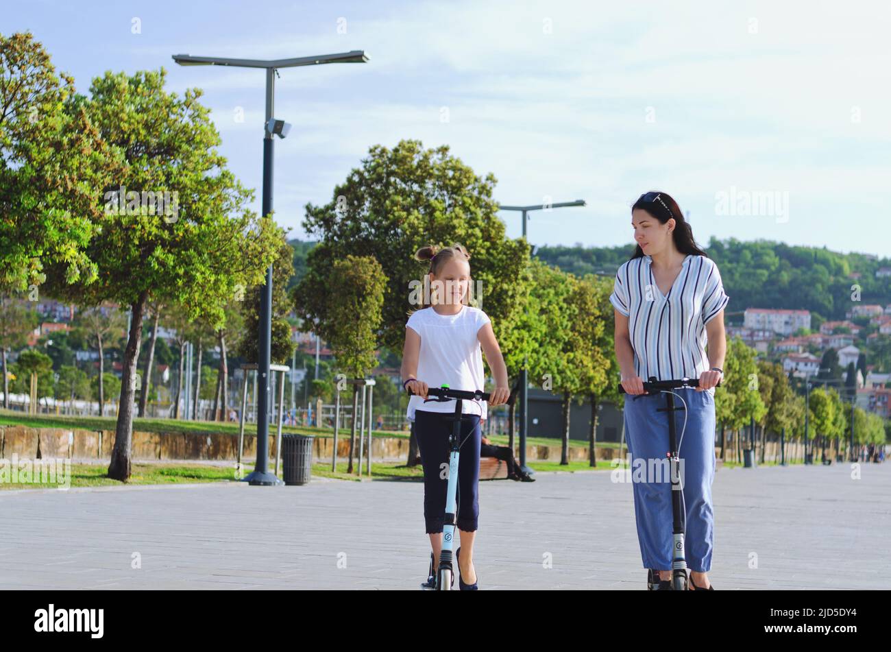 Happy family lifestyle and holiday concept. Mother, little girl riding scooters, walking in old city, street. Laughing on a summer sunny day Stock Photo