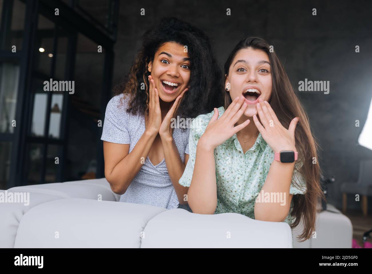 Two girls sit on the back of the sofa, look at the cameras and smile ...