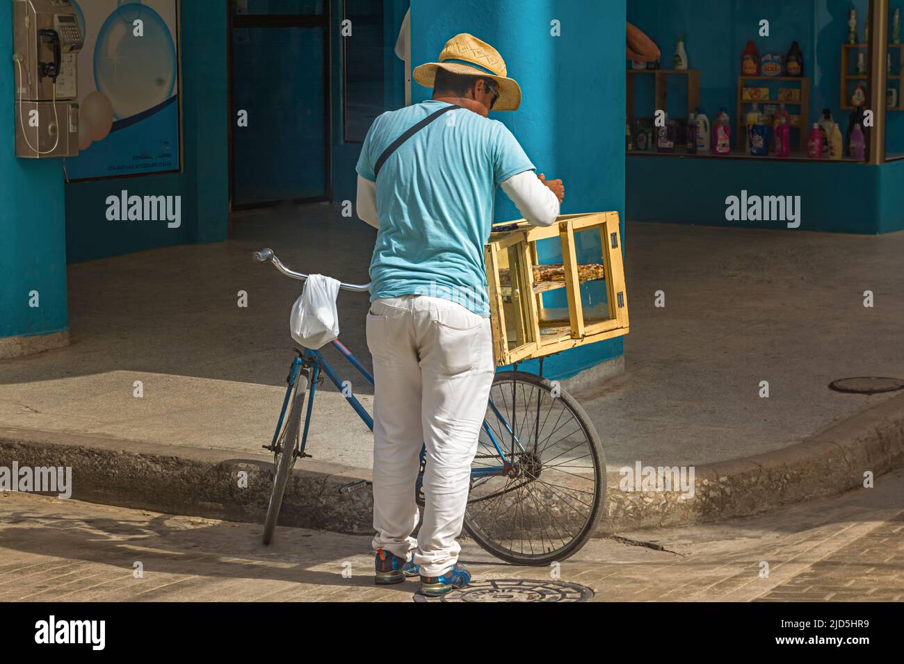 street vendor with a straw hat, white trousers and light blue T-Shirt and a bicycle sells bakery items in the town of Camagüey, Cuba Stock Photo