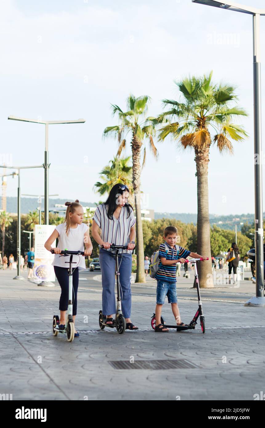 Happy family lifestyle and holiday concept. Mother, little boy, girl , riding scooters, walking in old city, street. Laughing on a summer sunny day. Stock Photo