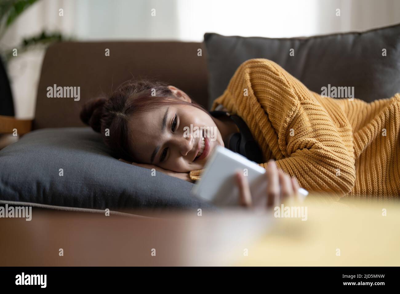 Happy young asian woman lying on sofa at living room and using mobile phone at home Stock Photo