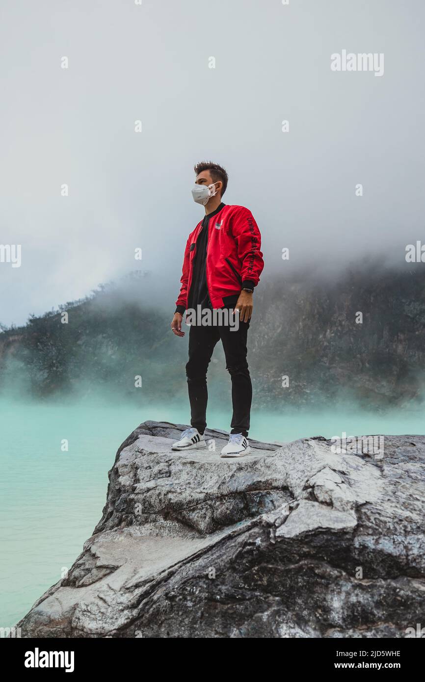 young man in red bomber jacket standing on rock at kawah putih sulfer lake Stock Photo