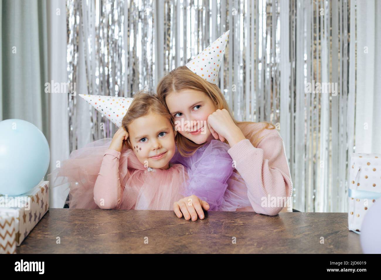 Portrait of two charming sisters celebrating their birthday. Girls in party hats and festive dresses sit same way, with their cheeks propped up with Stock Photo