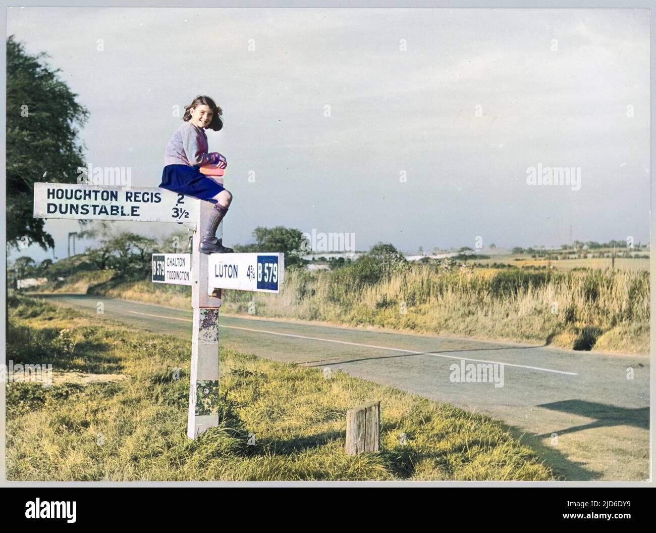 A girl in sturdy shoes and long socks, probably school uniform, perches on top of a signpost by the B579 road near Chalton in Bedfordshire. Colourised version of : 10237747       Date: 1950 Stock Photo