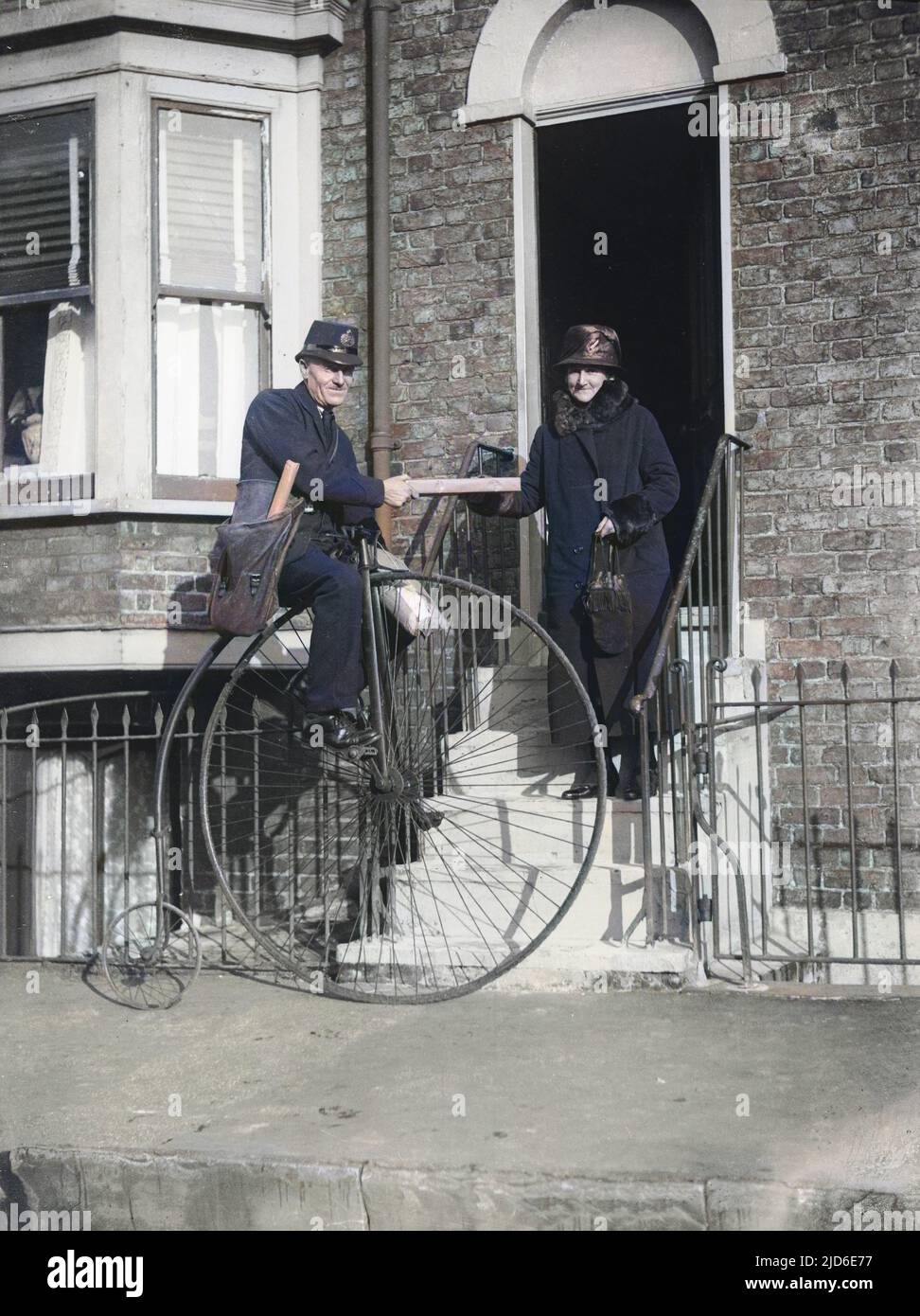 A postman on a penny farthing delivers a parcel to a woman dressed in a long coat with fur trim Colourised version of : 10092099       Date: 1930 Stock Photo
