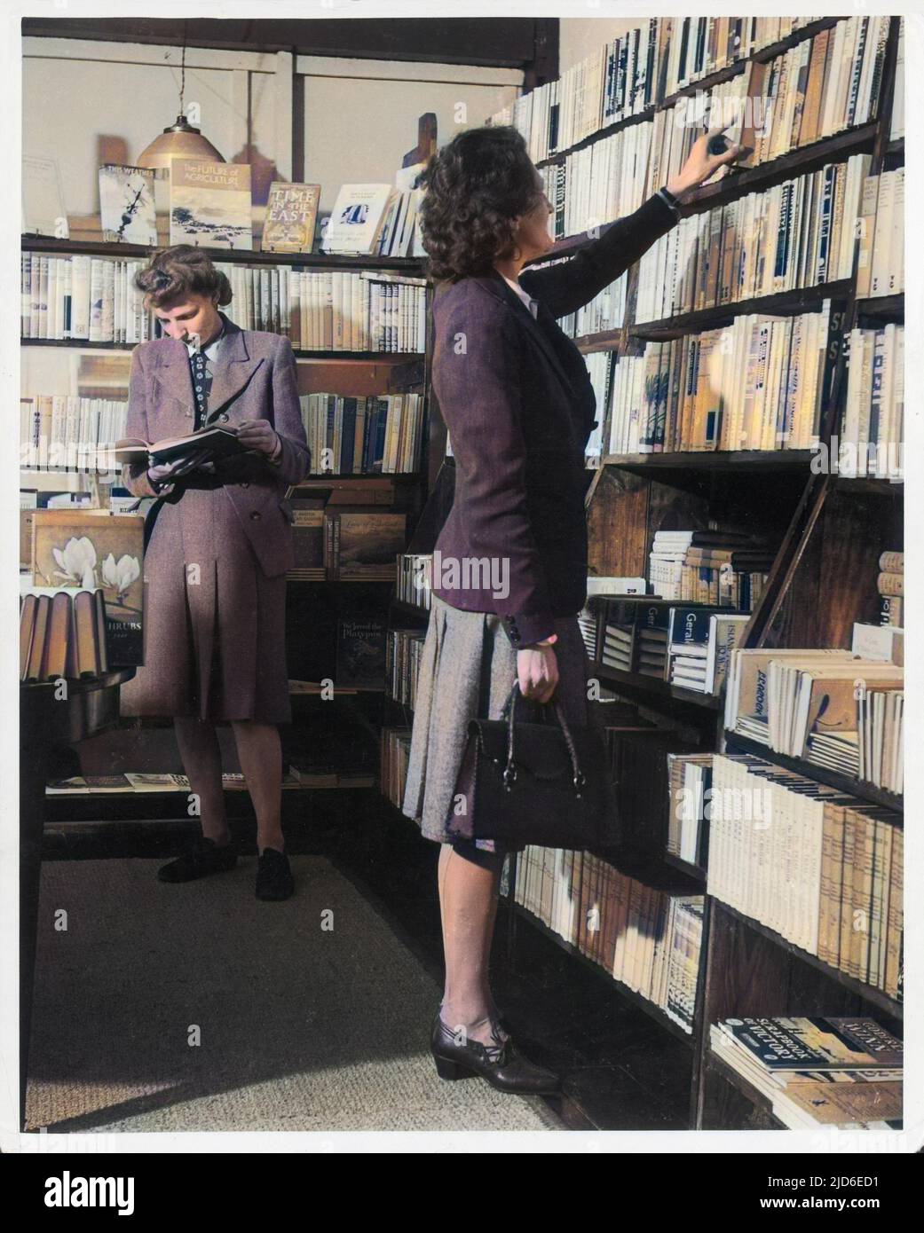 Two women, both wearing box pleated skirts, browse around a bookshop, which is selling the latest books, such as Godfrey Winn's 'Scrapbook of Victory'. Colourised version of : 10144388       Date: late 1940s Stock Photo