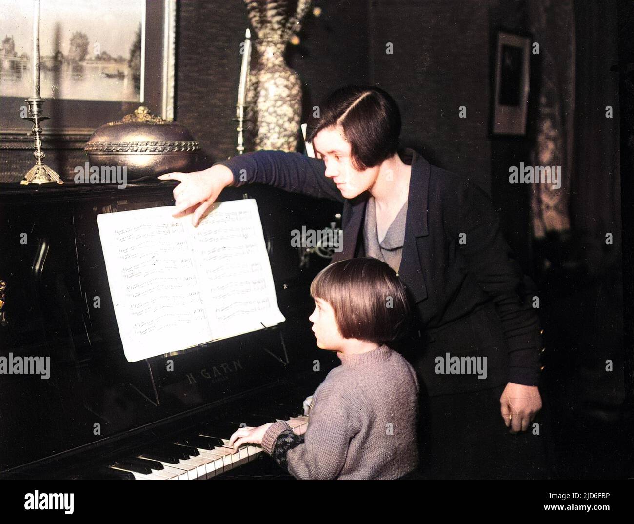A female piano teacher turns over the pages of the sheet music as little girl pupil plays the piece. Colourised version of : 10164308       Date: early 1930s Stock Photo