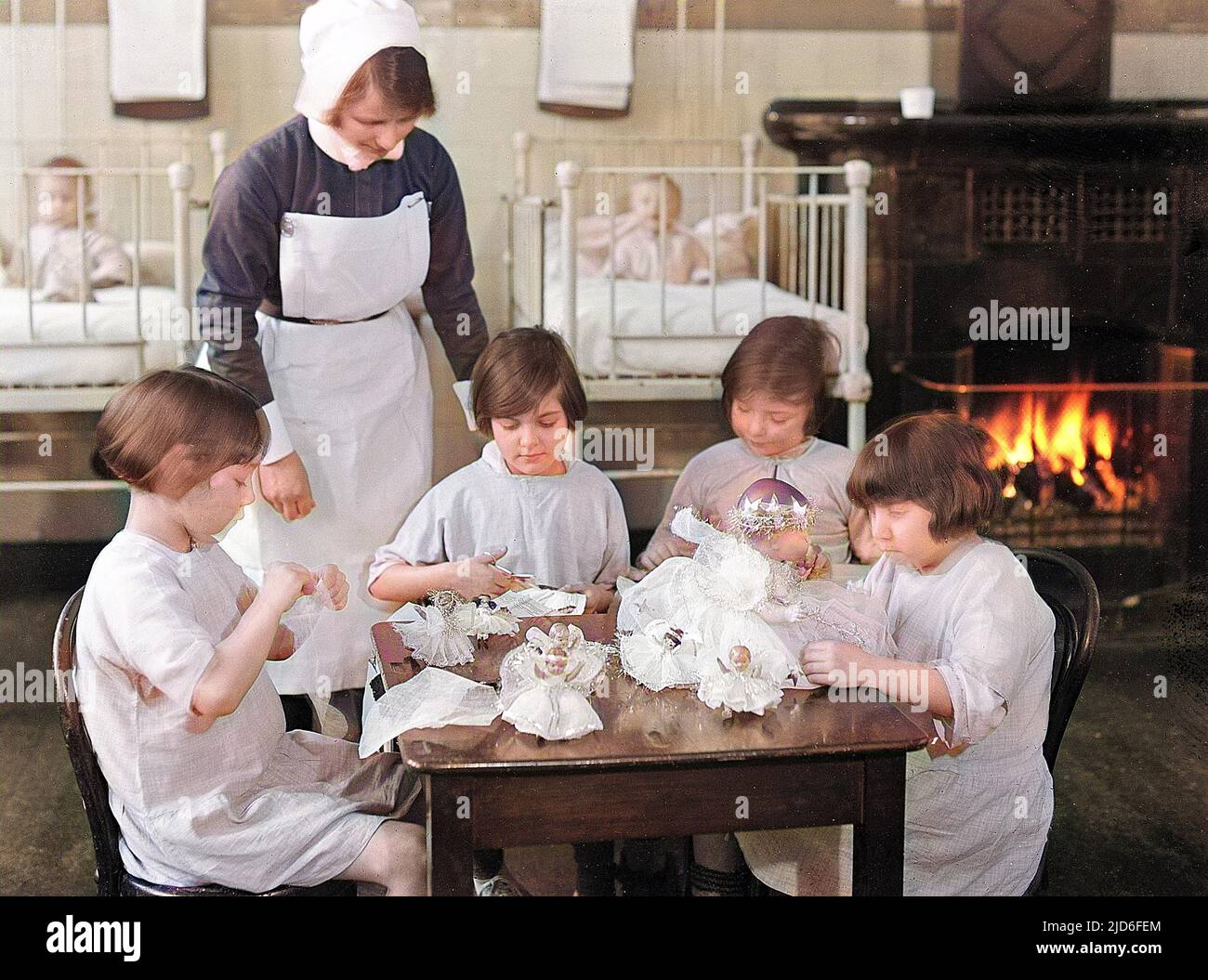 Sick little girls playing with dolls in the children's ward, supervised by a kindly nurse. Behind them are babies in cots and a roaring fire. Colourised version of : 10164424       Date: early 1930s Stock Photo