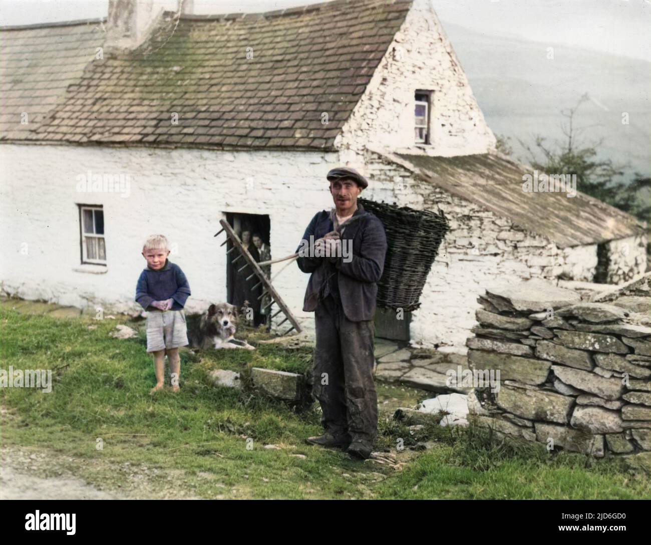 A man delivering peat in a basket to a typical Irish cottage, watched by a little boy and two women in the doorway. Colourised version of : 10171057       Date: Oct-38 Stock Photo