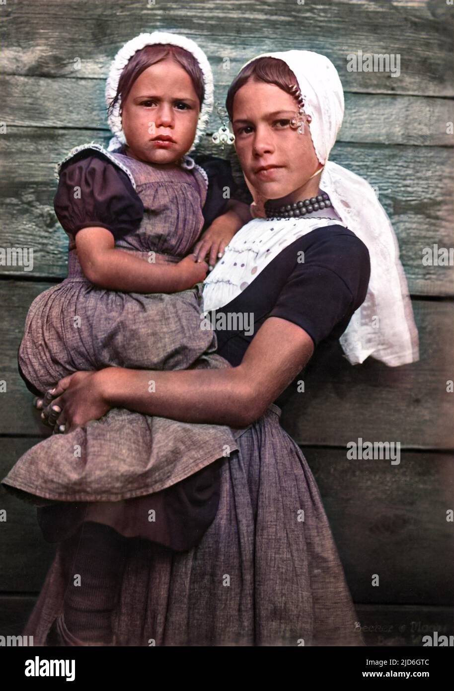 Two Dutch girls, probably sisters, in traditional costume, the older girl carrying the younger girl in her arms. Colourised version of : 10182678       Date: 1930s Stock Photo