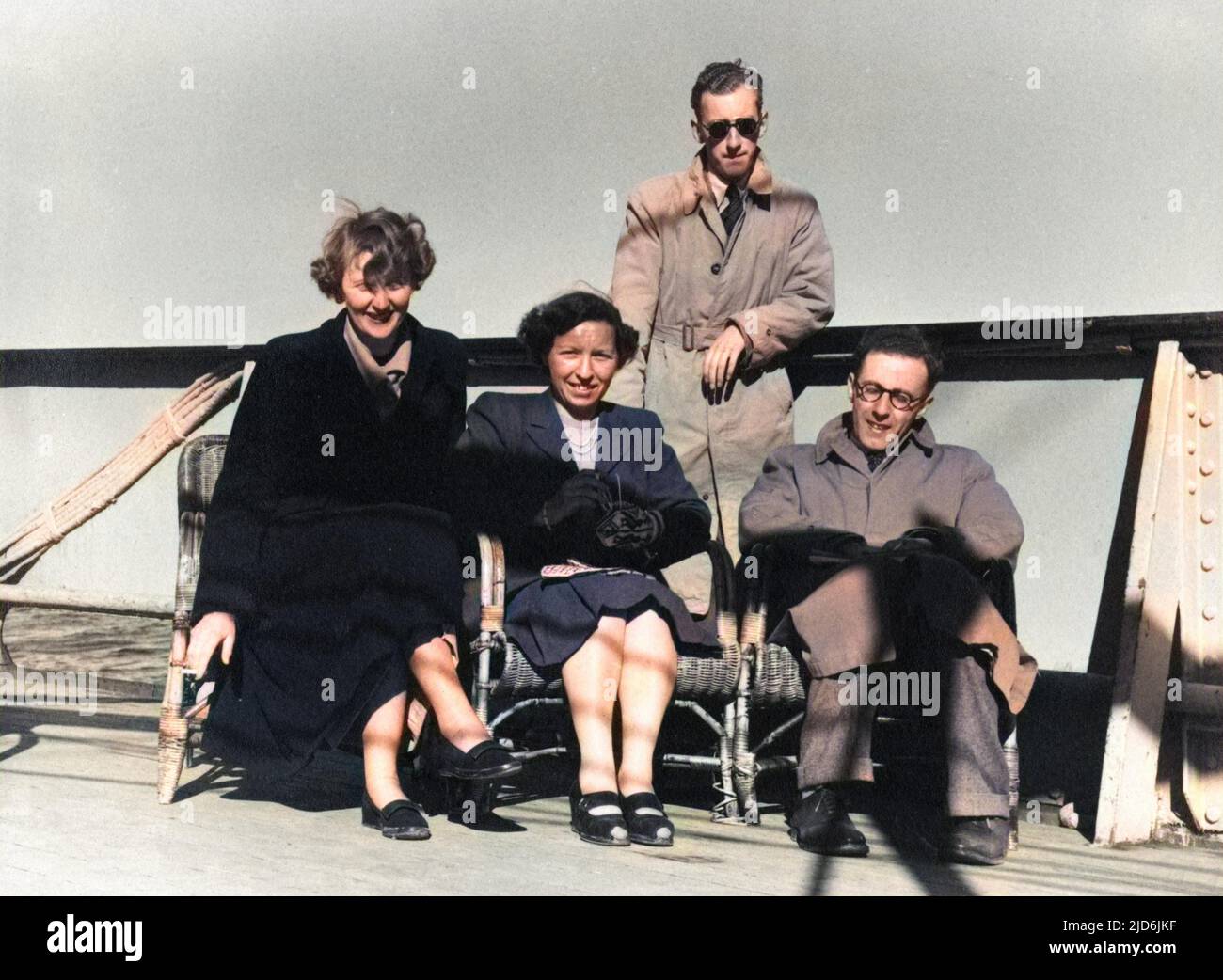 Slightly windswept passengers on deck aboard the ferry on the way to a Holiday in Norway. Sitting on wicker chairs close to the rail, wrapped up in raincoats and mittens! Colourised version of: 10794428       Date: early 1950s Stock Photo