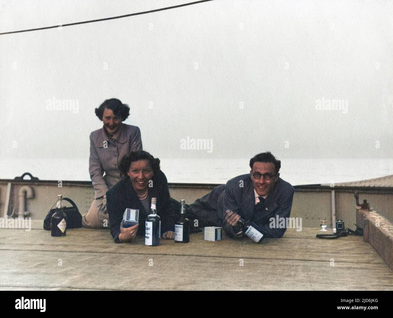 Two women and a man pose on the deck of a Norwegian ferry boat wit their duty free spirits and cigarettes. Following WW2 there was still considerable rationing of food and drink in the UK, hence the advantages of purchases on an overseas trip. Colourised version of: 10794431       Date: early 1950s Stock Photo