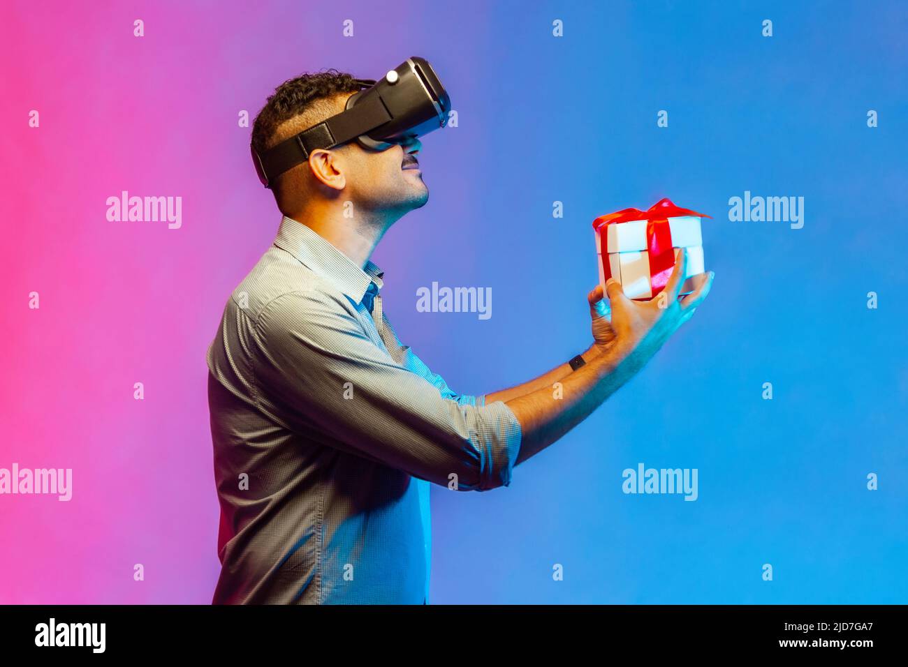 Side view of man in VR virtual reality glasses holding wrapped gift box and smiling joyfully, giving present, celebrating holiday. Indoor studio shot isolated on colorful neon light background. Stock Photo