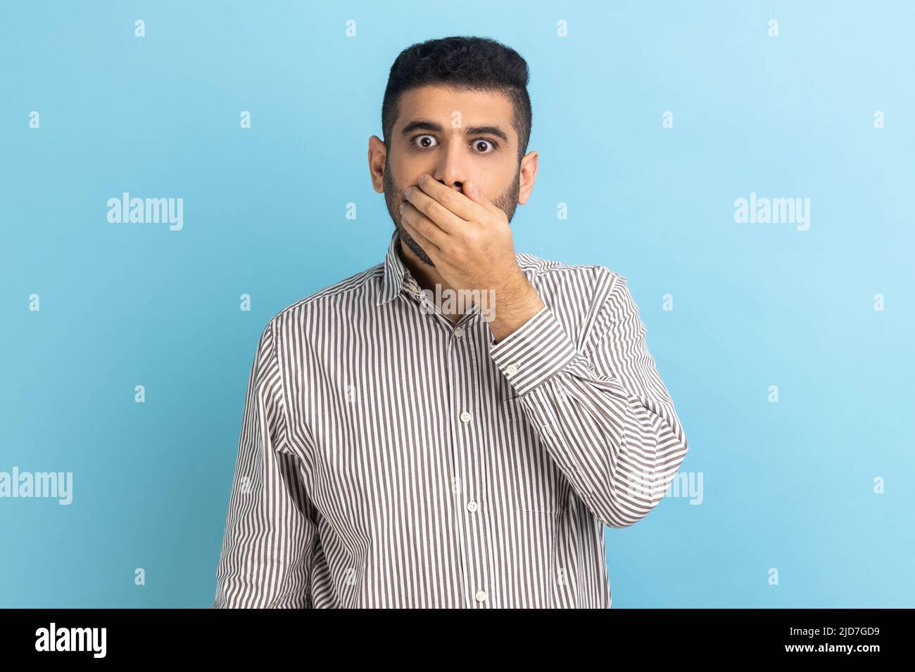 I won't tell. Portrait of amazed businessman covering mouth with hand, keeping secret, terrified with shocking news, wearing striped shirt. Indoor studio shot isolated on blue background. Stock Photo