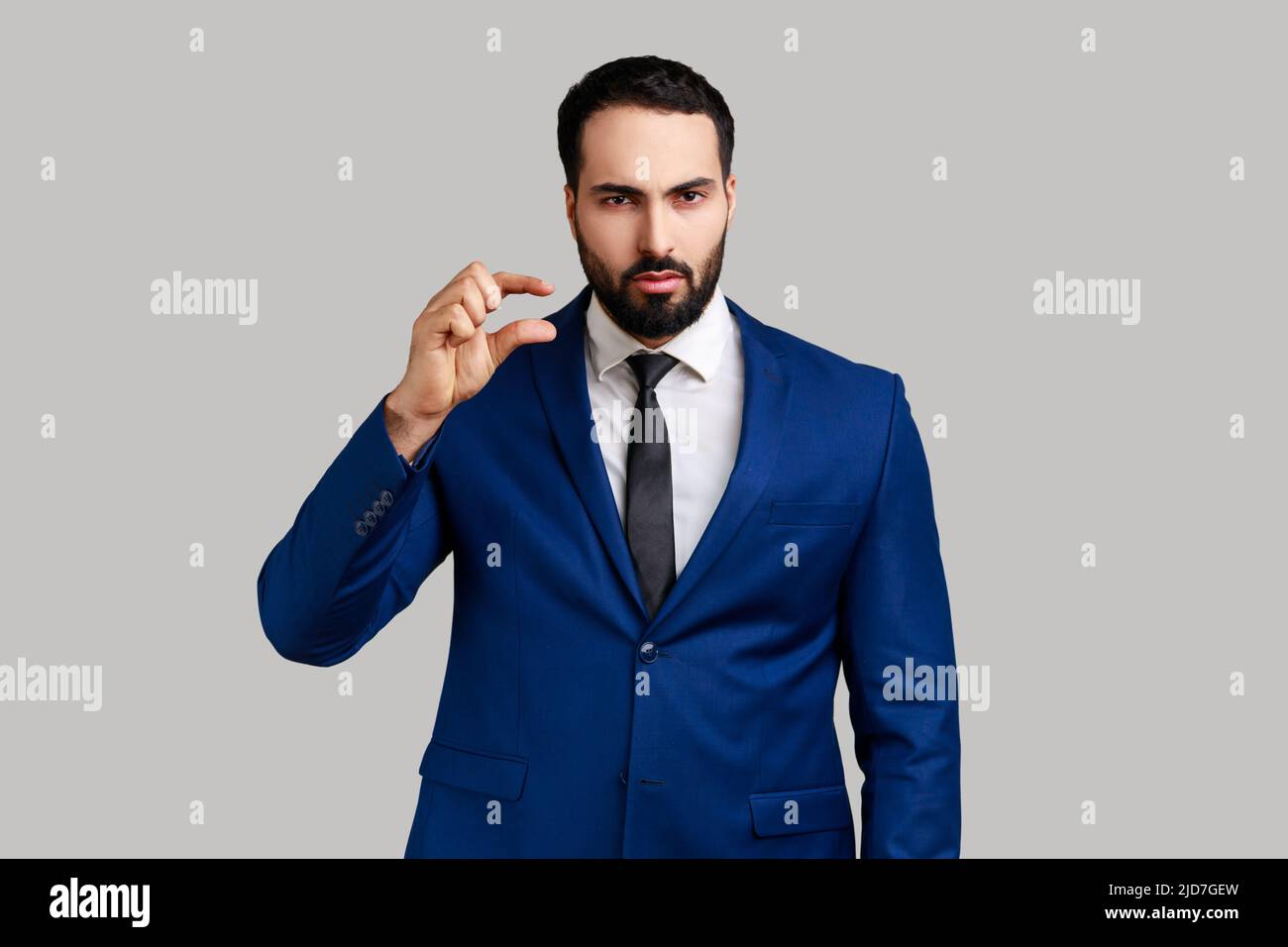 Bearded man looking disappointed and showing a little bit gesture, small amount or low scale sign, few centimeters, wearing official style suit. Indoor studio shot isolated on gray background. Stock Photo