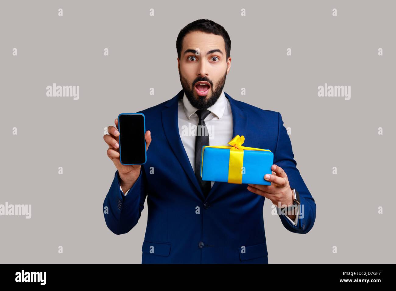Shocked businessman holding gift box and cell phone with empty display for online shopping advertising, wearing official style suit. Indoor studio shot isolated on gray background. Stock Photo