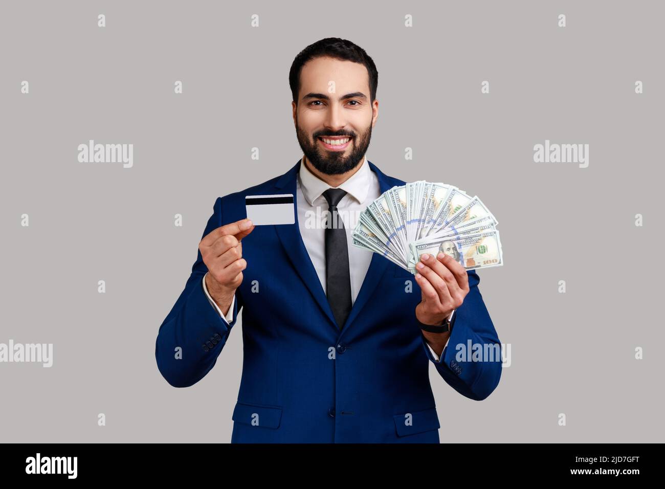 Portrait of smiling bearded businessman holding credit card and lot of money, being glad by cash deposits, wearing official style suit. Indoor studio shot isolated on gray background. Stock Photo