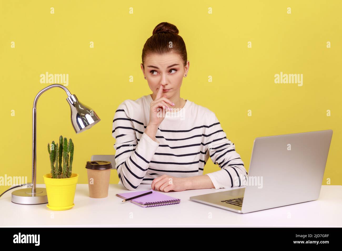Lazy woman employee wearing striped shirt picking nose and looking away with comical expression, having break at workplace. Indoor studio studio shot isolated on yellow background. Stock Photo