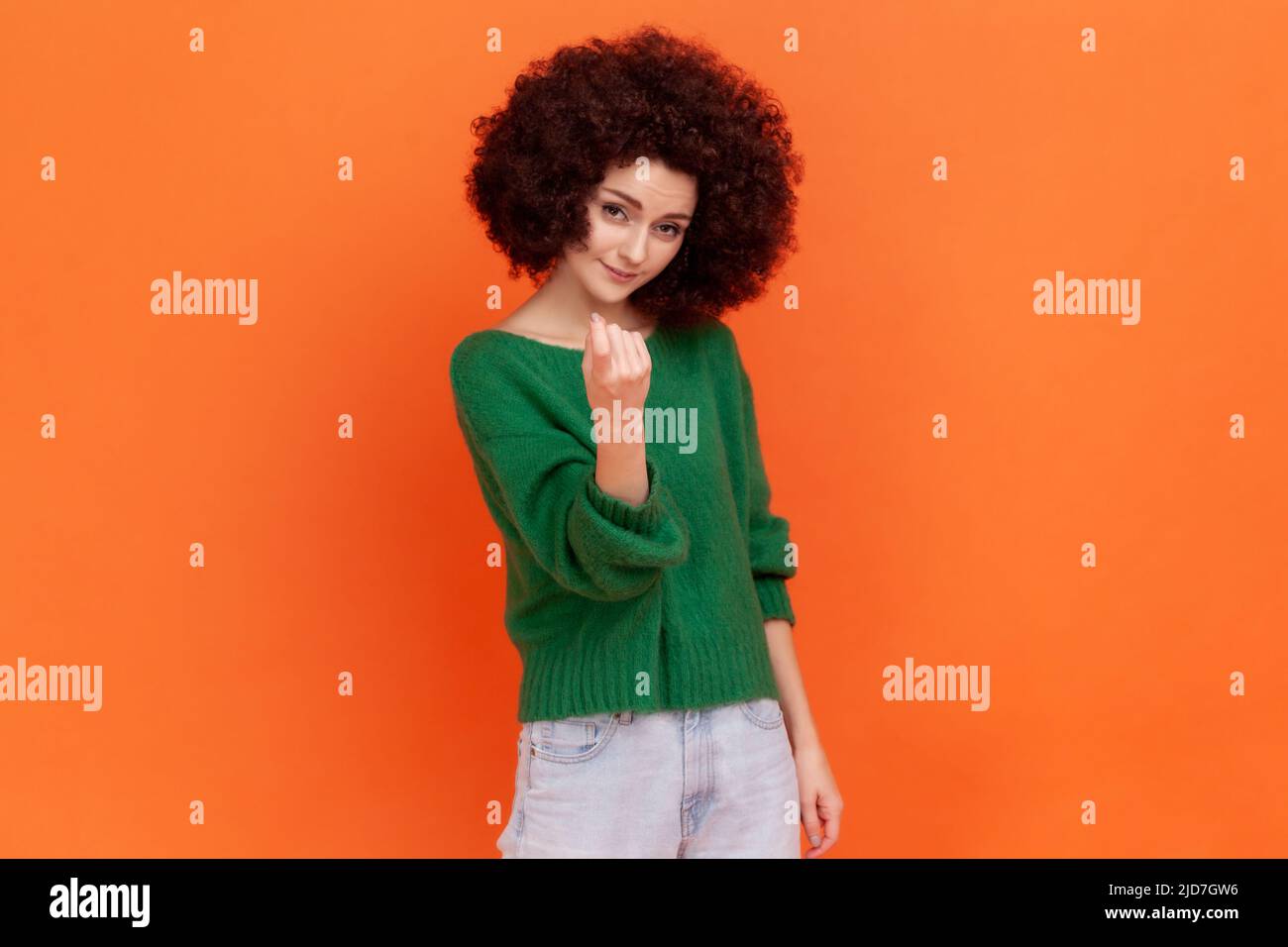 Come here. Woman with Afro hairstyle wearing green casual style sweater making beckoning gesture, inviting to come, flirting and looking playful. Indoor studio shot isolated on orange background. Stock Photo