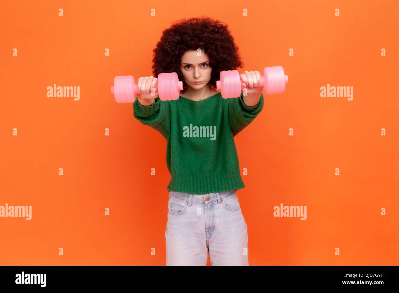 Strong confident woman with Afro hairstyle wearing green casual style sweater holding out pink dumbbells to camera, work out alone. Indoor studio shot isolated on orange background. Stock Photo